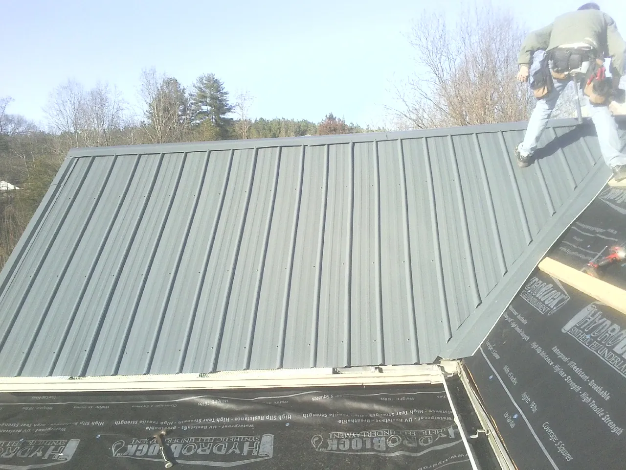 Crew installing dark green standing seam panels over synthetic underlayment in Farnhamville