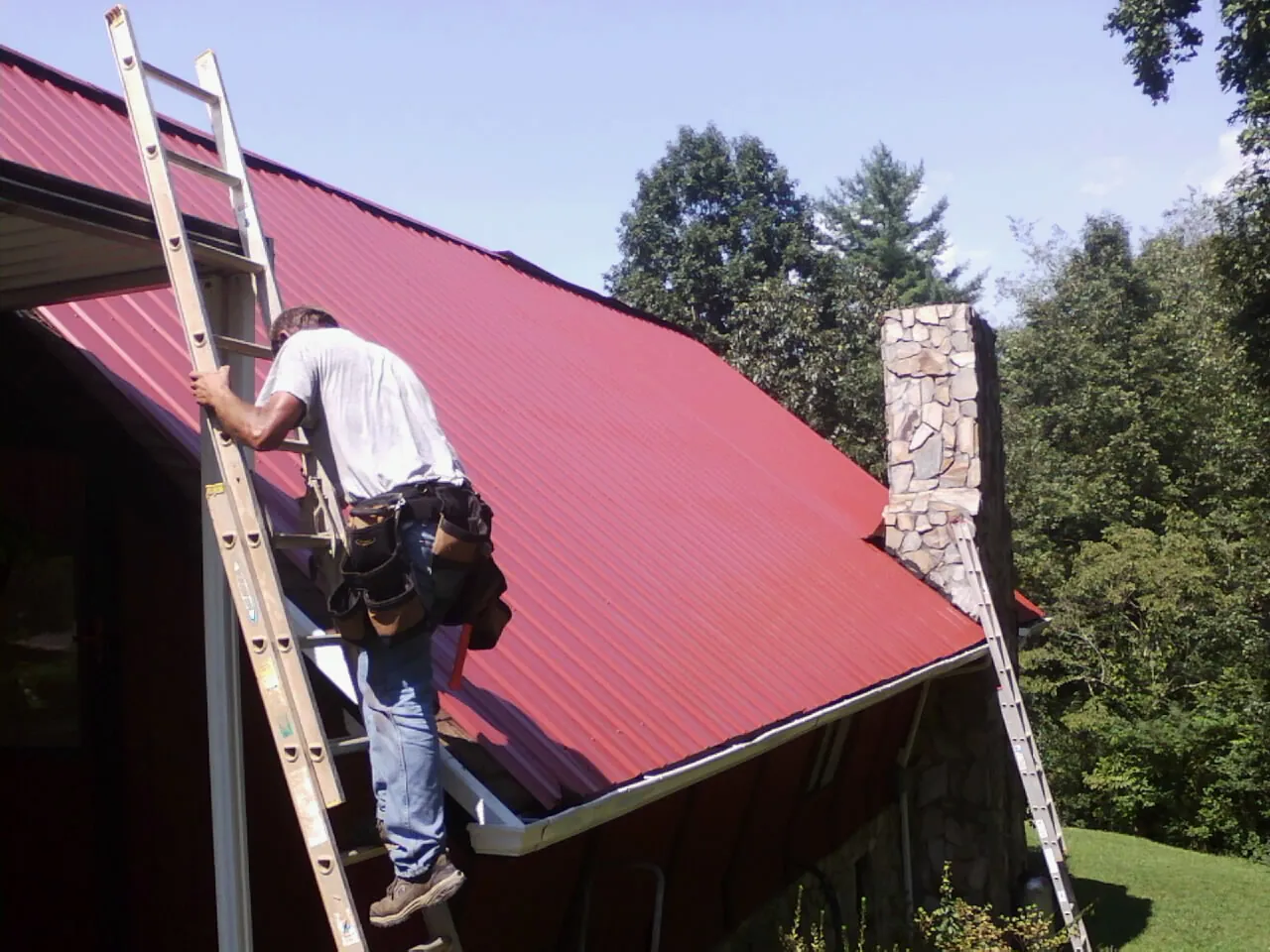 Roofer on a ladder beside a red metal roof and stone chimney — Soffit & Fascia Repair by Roofing Brothers in Farnhamville
